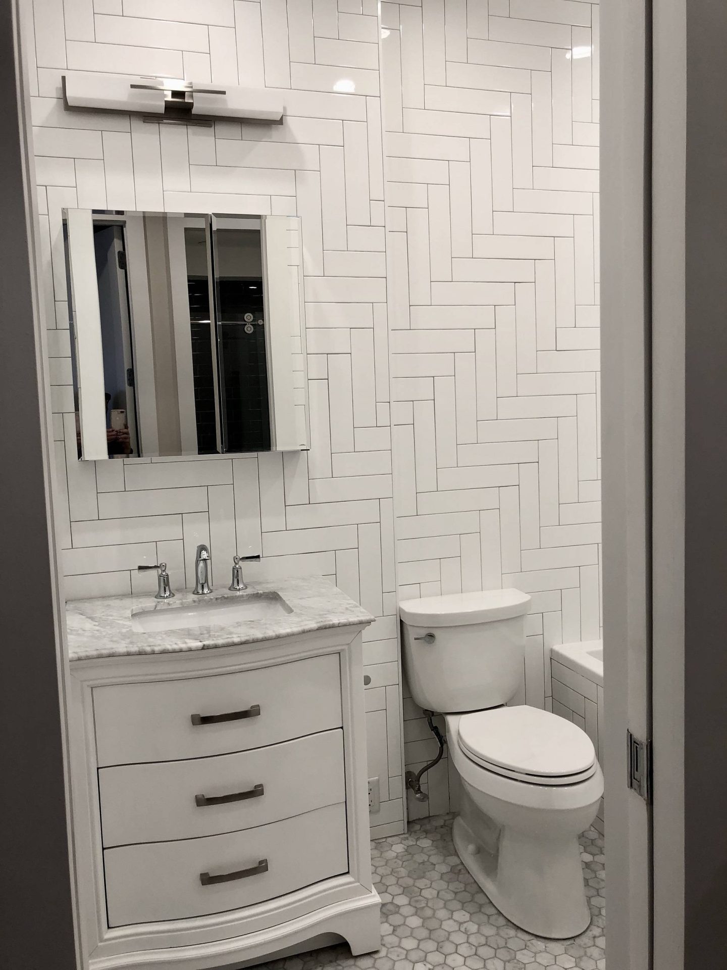 A small bathroom with white herringbone wall tiles, a vanity with a marble countertop, a toilet, a mirrored medicine cabinet, and hexagonal floor tiles.
