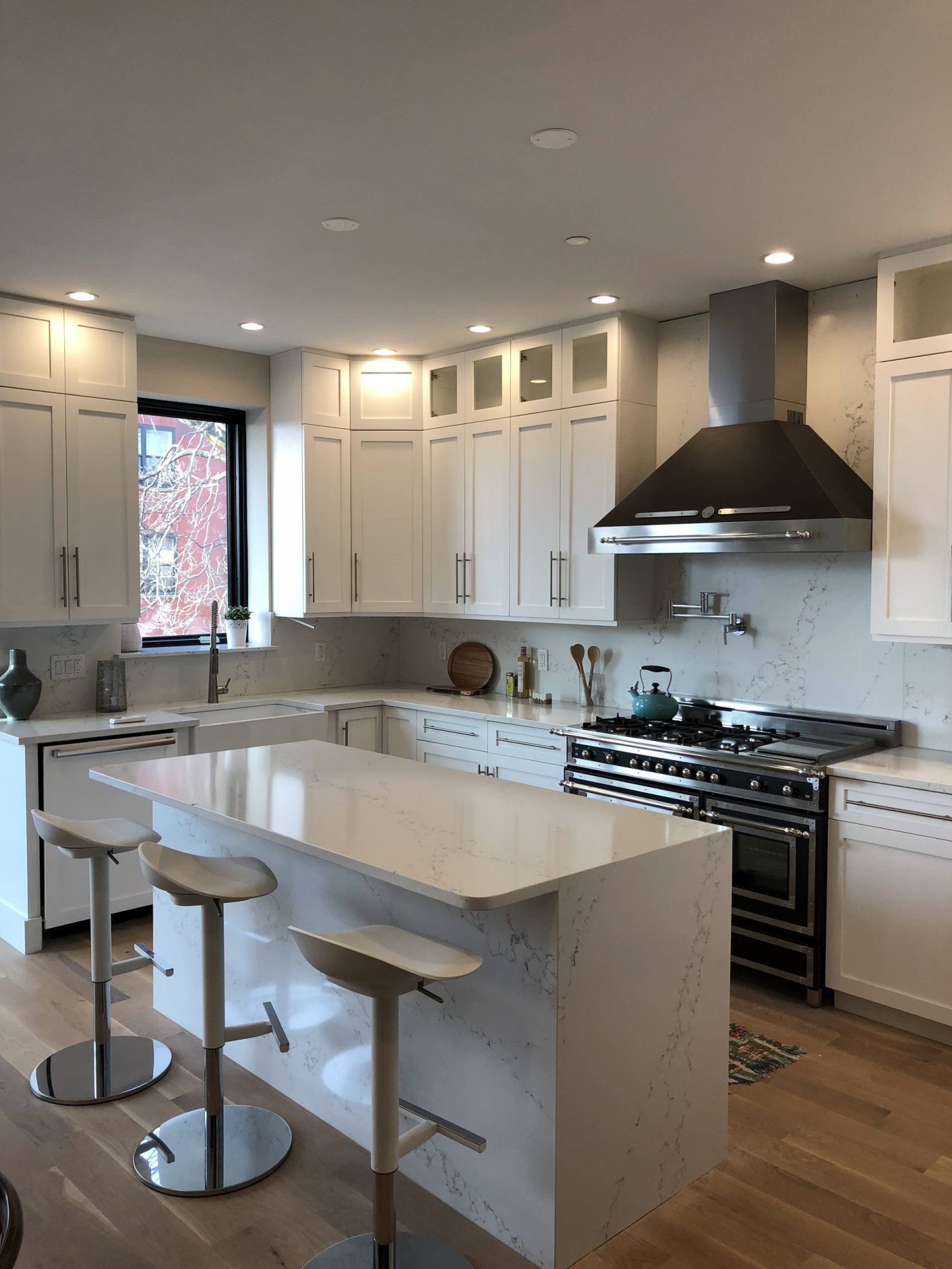 Modern kitchen with white cabinets, marble countertops, stainless steel appliances, a large range hood, and three white bar stools at an island on a light wood floor.