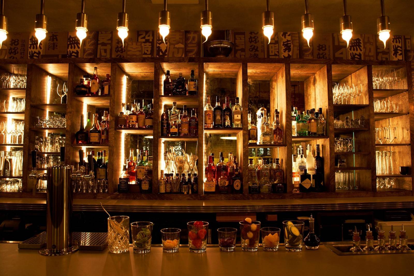 A well-lit bar with shelves of liquor bottles and glasses, with various cocktail ingredients and garnishes arranged in glasses on the counter.