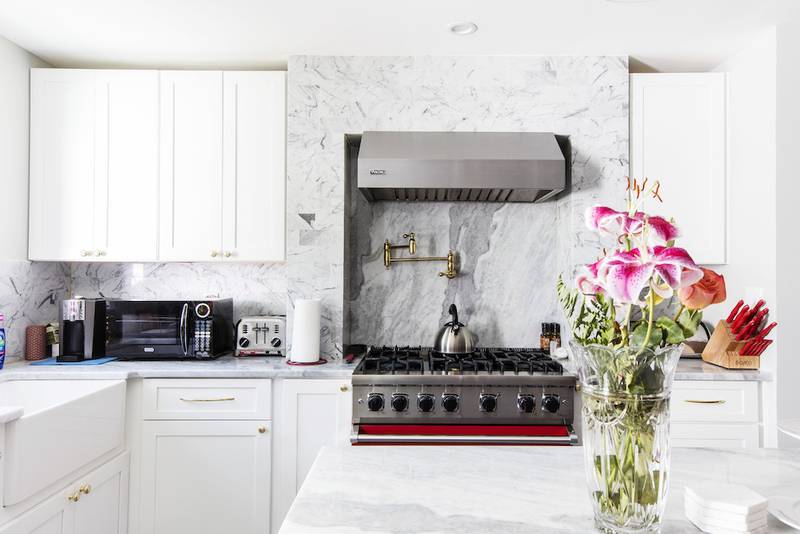 Modern kitchen with white cabinets, marble countertops and backsplash, stainless steel stove with range hood, small appliances, and a vase of pink flowers on the counter.