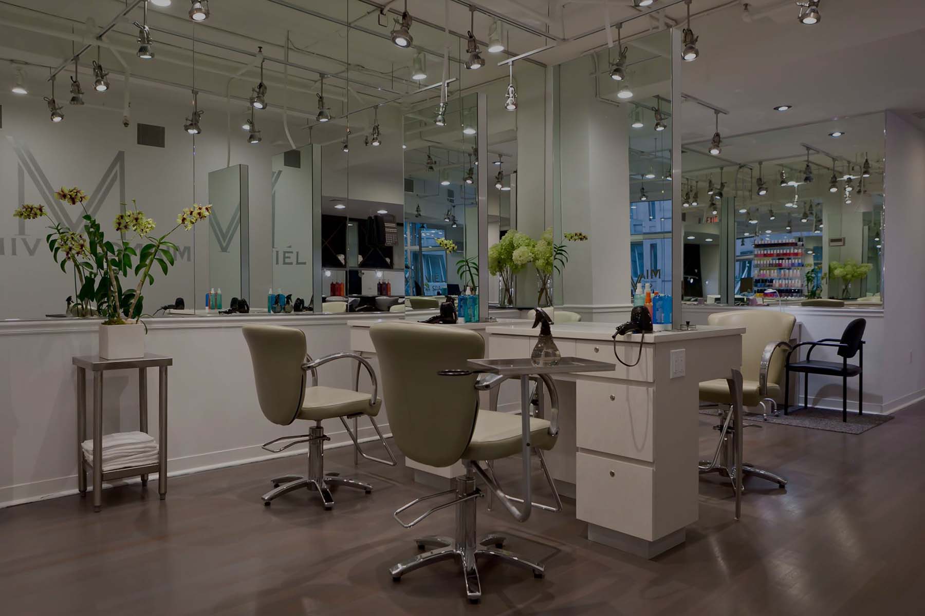 Modern hair salon interior with styling chairs, mirrors, desks, and plants; overhead track lighting and reflection of equipment in the mirrors.