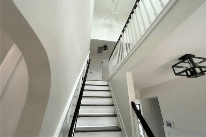 View looking up a white stairwell with dark handrails, leading to a landing and hallway above, with a ceiling fan and modern light fixtures visible.